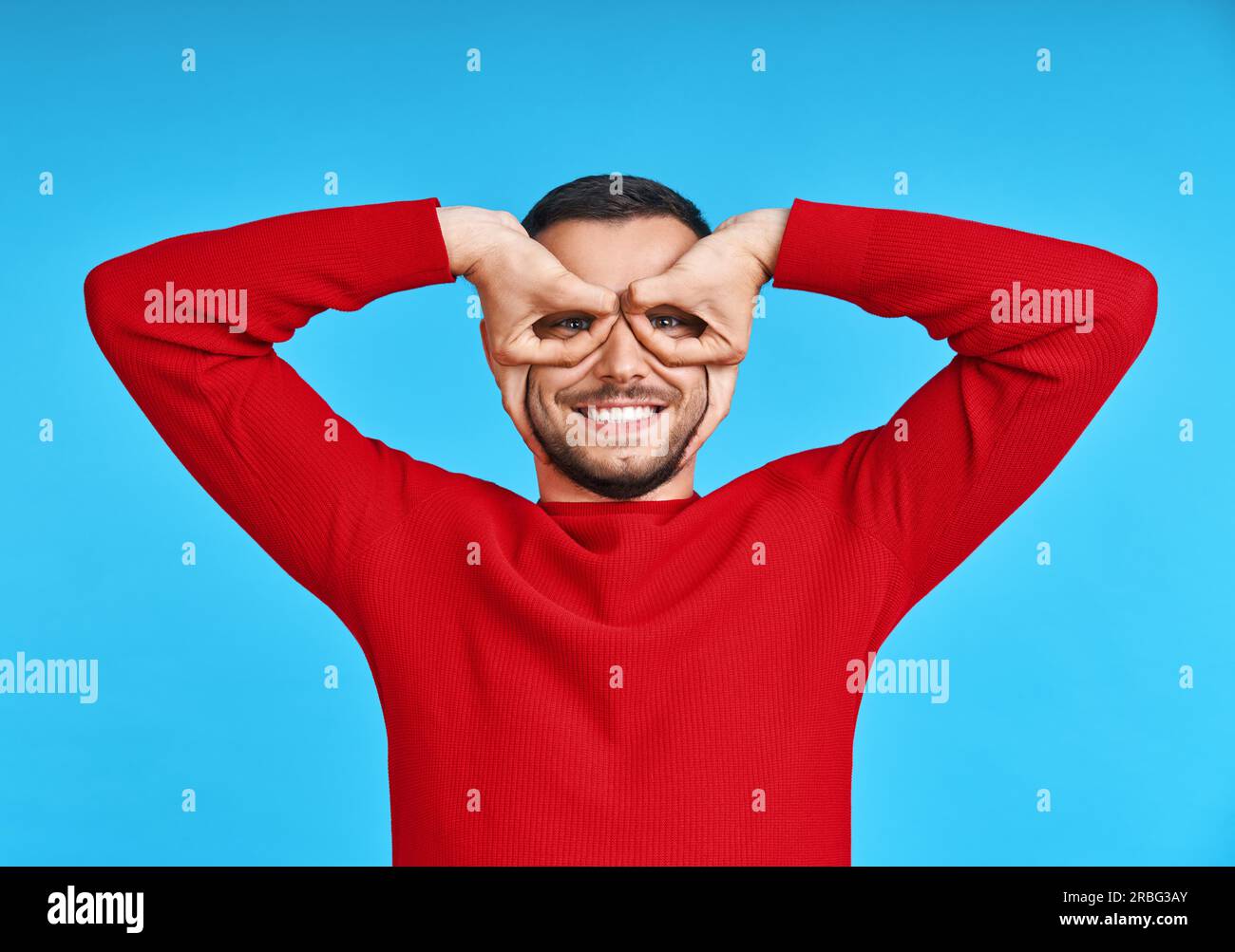 Ritratto di felice uomo creativo rendendo gli occhiali del dito su blu di sfondo per studio. Divertimento e concetto di persone Foto Stock