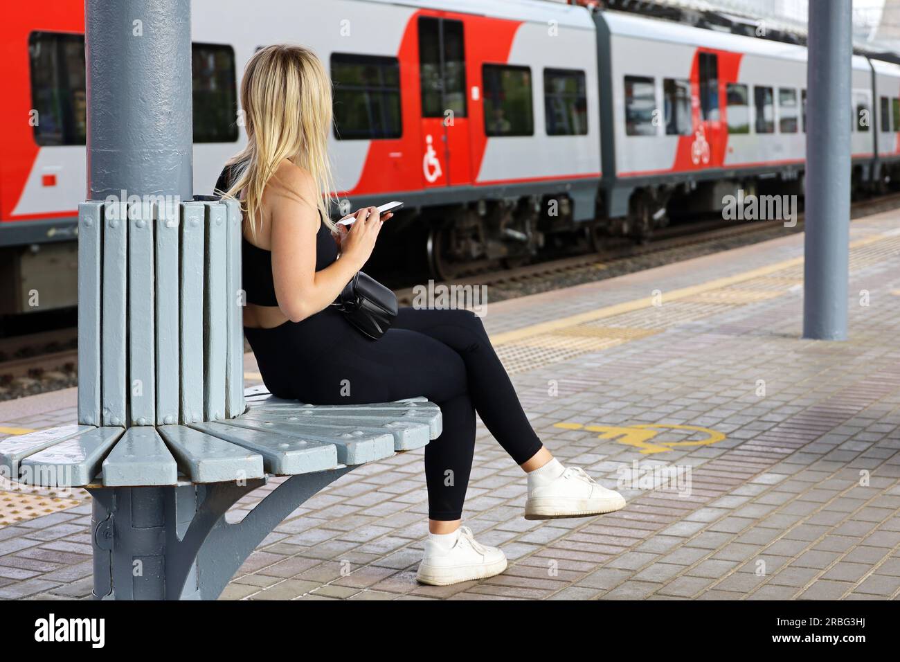 Ragazza bionda in attesa del treno in arrivo seduta con lo smartphone in mano sulla stazione ferroviaria. Passeggeri del treno pendolare, viaggi in estate Foto Stock