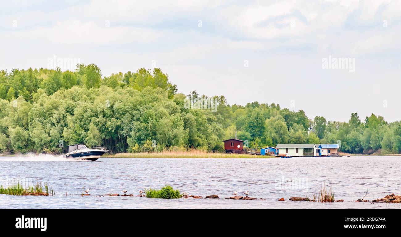 Piccola casa vicino al fiume Dnieper a Kiev, Ucraina, all'inizio della primavera, sotto un cielo nuvoloso. Un bianco e nero speedboat passa davanti Foto Stock
