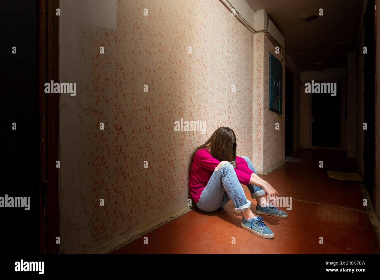 Una donna triste e disperata siede in un corridoio buio illuminato da una luce cupa. Il suo dolore e i suoi molti problemi l'hanno spinta in completo isolamento. Foto Stock