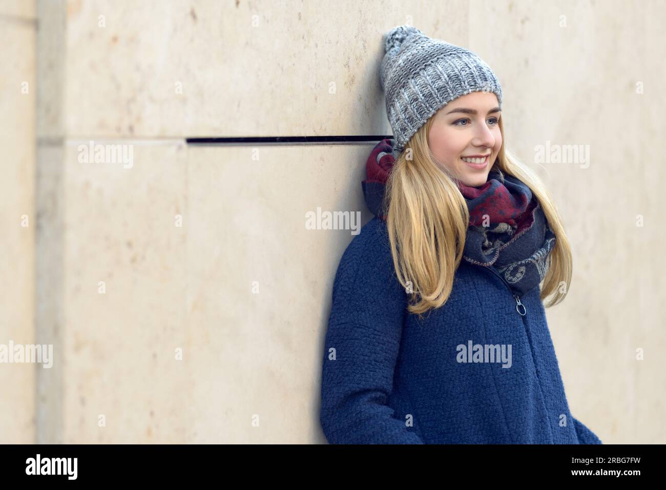 Sorridente attraente giovane donna bionda in un caldo inverno blu permanente outfit fantasticando appoggiato contro una parete esterna fissando in distanza Foto Stock