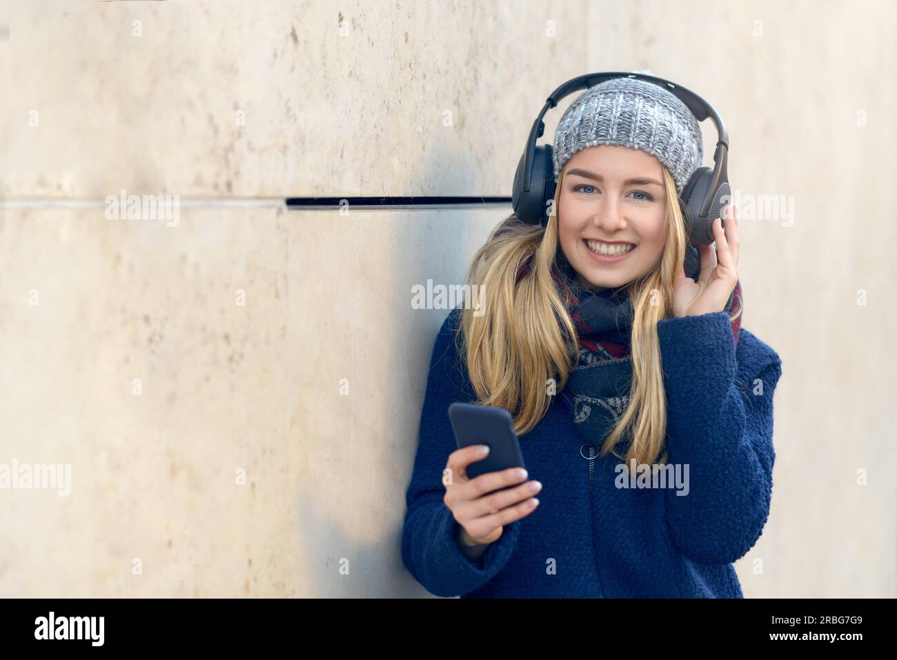 Giovane donna bionda con cappotto e cappello in piedi all'esterno contro una parete di cemento tenendo lo smartphone in mano con gli occhi chiusi sorridendo ascoltando la musica Foto Stock