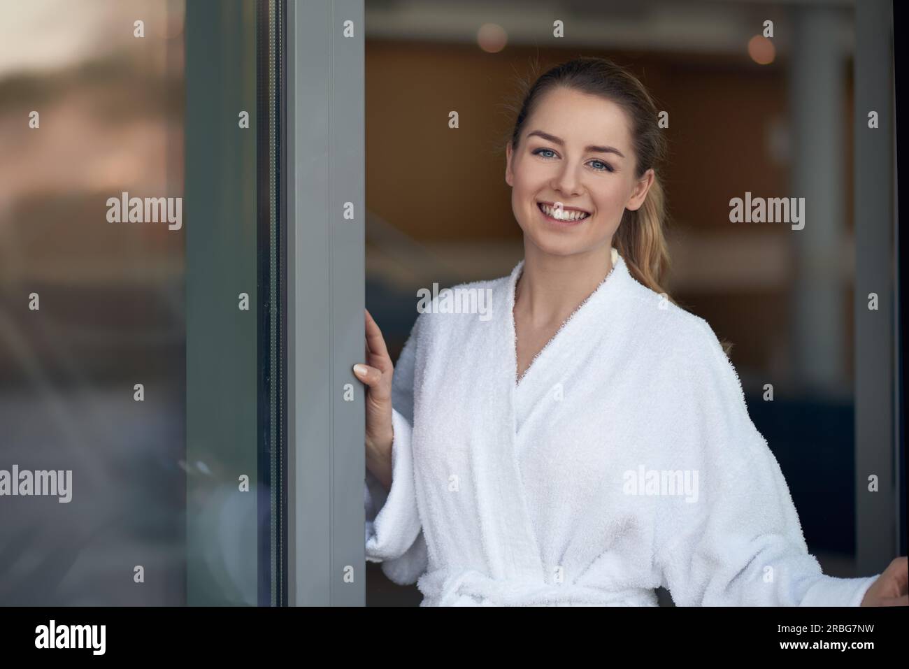 Felice giovane donna in accappatoio bianco apertura di una porta di vetro e sorridente alla telecamera, con spazio di copia Foto Stock