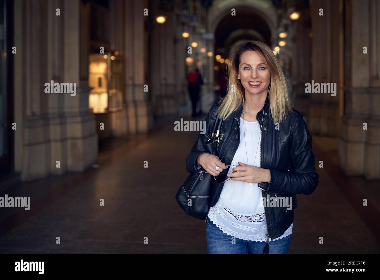 Attraente trendy giovane donna a piedi attraverso un porticato con la borsetta sulla sua spalla sorridendo felicemente alla fotocamera con spazio di copia Foto Stock