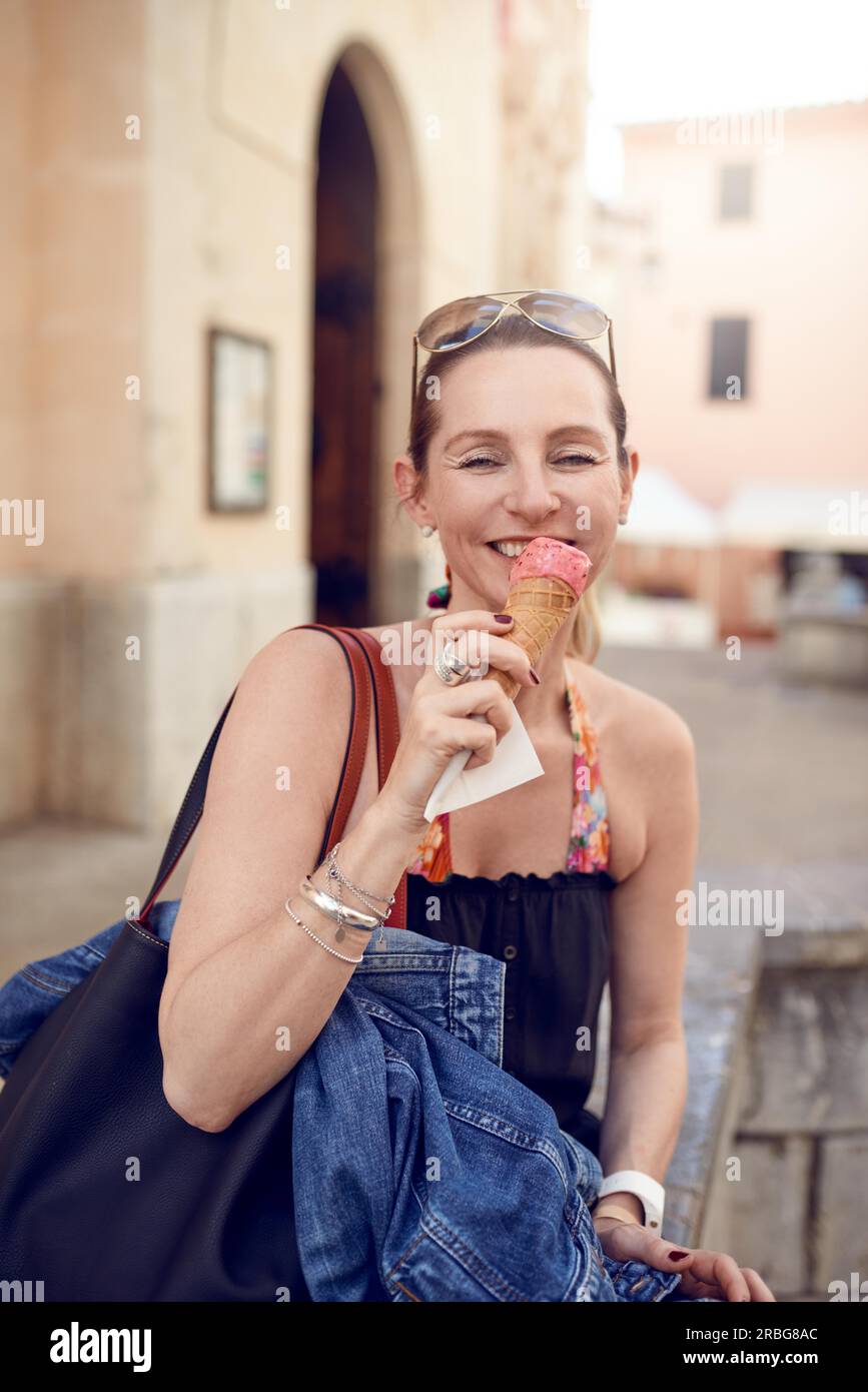 Sorridente attraente donna elegante con i suoi occhiali da sole sulla sua fronte godendo di un gelato in un cono esterno in un vicolo pedonale in una città Foto Stock