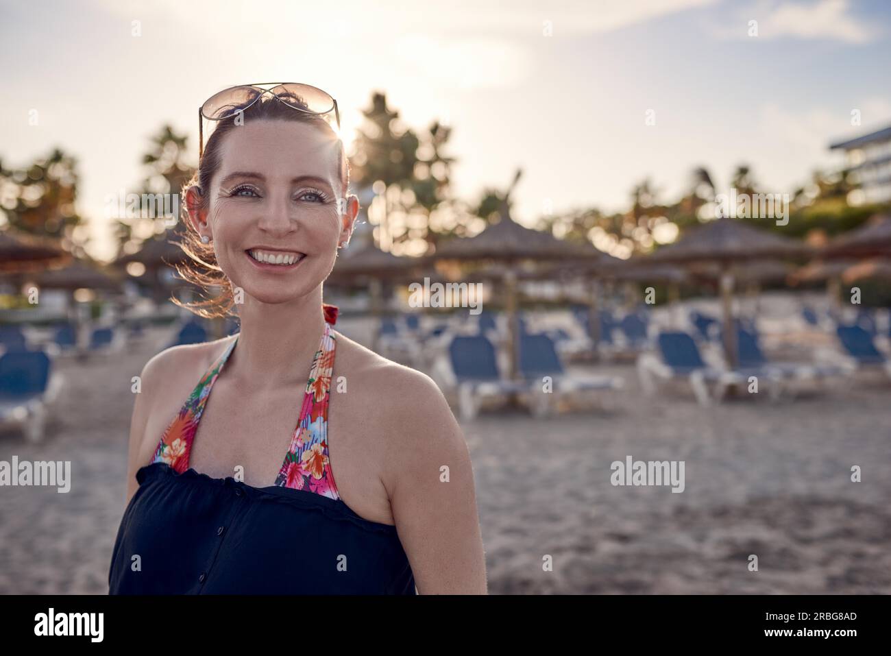 Bella, felice ed elegante donna sulla spiaggia al tramonto con sedie reclinabili dietro di lei sulla sabbia che guarda da parte con un sorriso vivace e incantevole su un Foto Stock