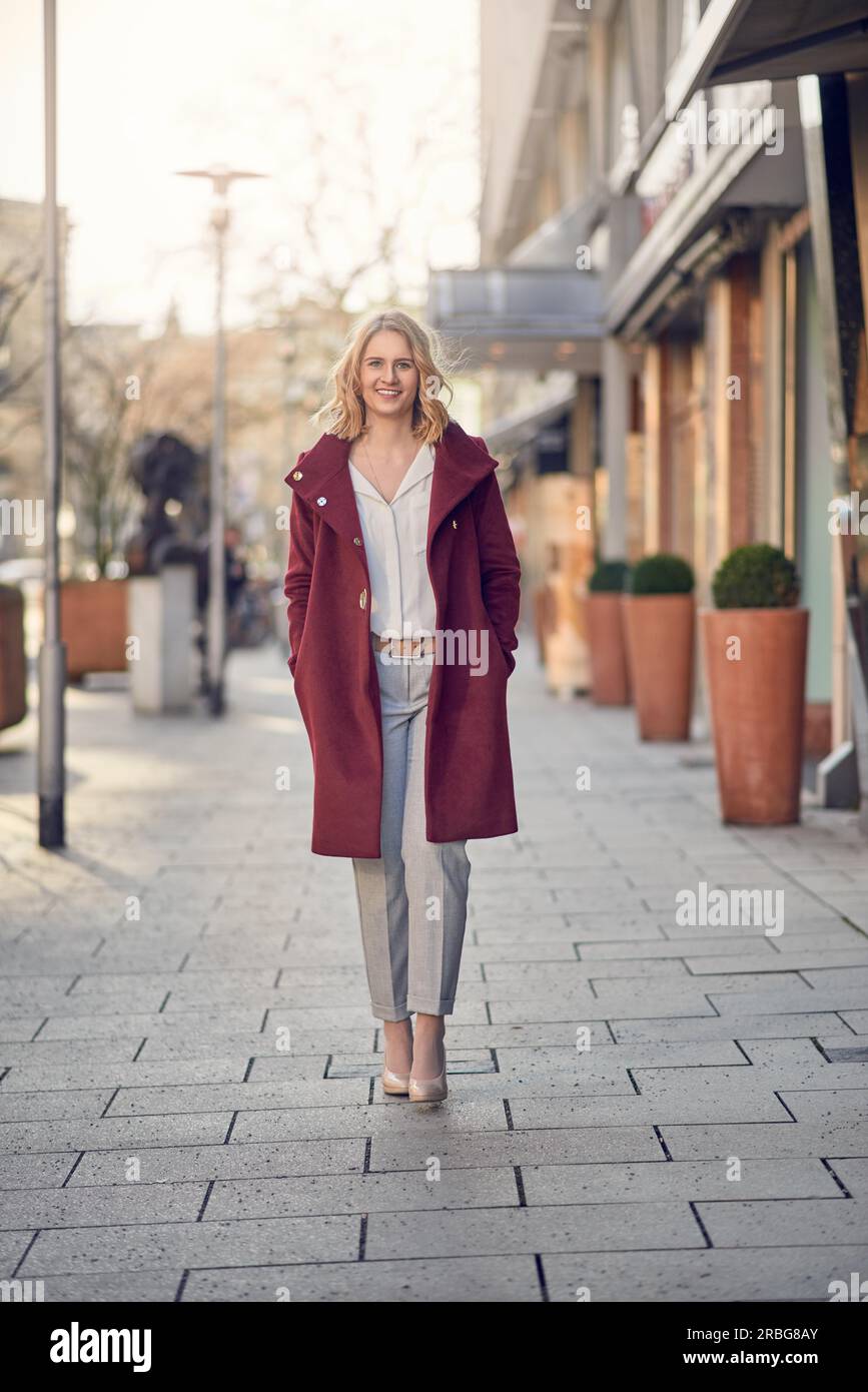 Elegante Attraente giovane donna in un marrone rossiccio soprabito in piedi con le mani nelle sue tasche in una strada di città di sorridere alla telecamera Foto Stock