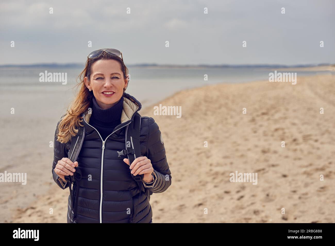 Donna attraente e sana che si gode un'escursione sulla spiaggia in una fredda giornata ricoperta di sole sorridendo davanti alla macchina fotografica sulla sabbia che le regge le cinghie Foto Stock