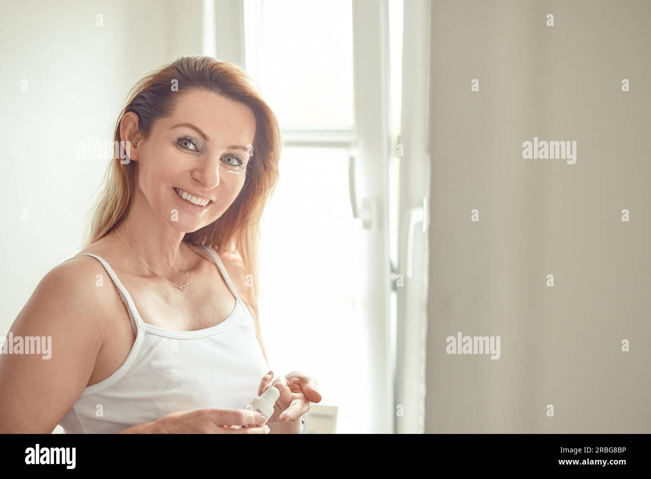 Bella donna bionda in jeans e maglietta bianca in piedi nel bagno di casa e l'applicazione di crema per il viso sotto i suoi occhi, guardando la fotocamera e sorridente Foto Stock