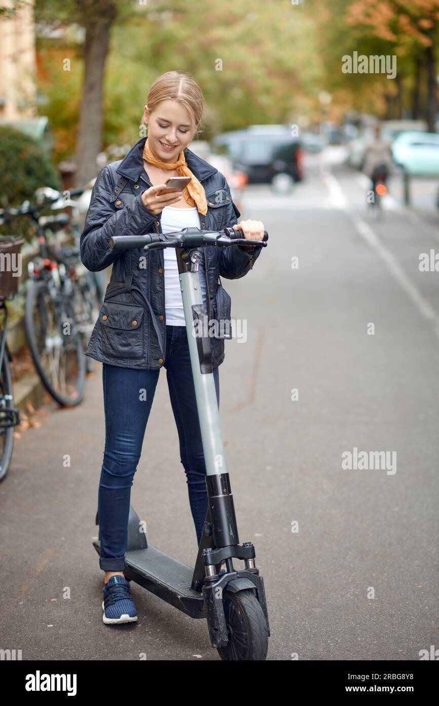 Giovane donna con scooter elettrico in strada, guarda il suo telefono e sorride. Servizio di noleggio di mezzi pubblici, moderno Foto Stock