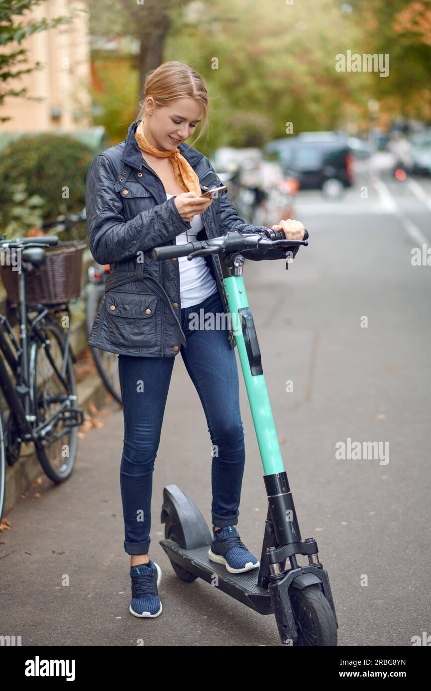 Giovane donna con scooter elettrico in strada, guarda il suo telefono e sorride. Servizio di noleggio di mezzi pubblici, moderno Foto Stock