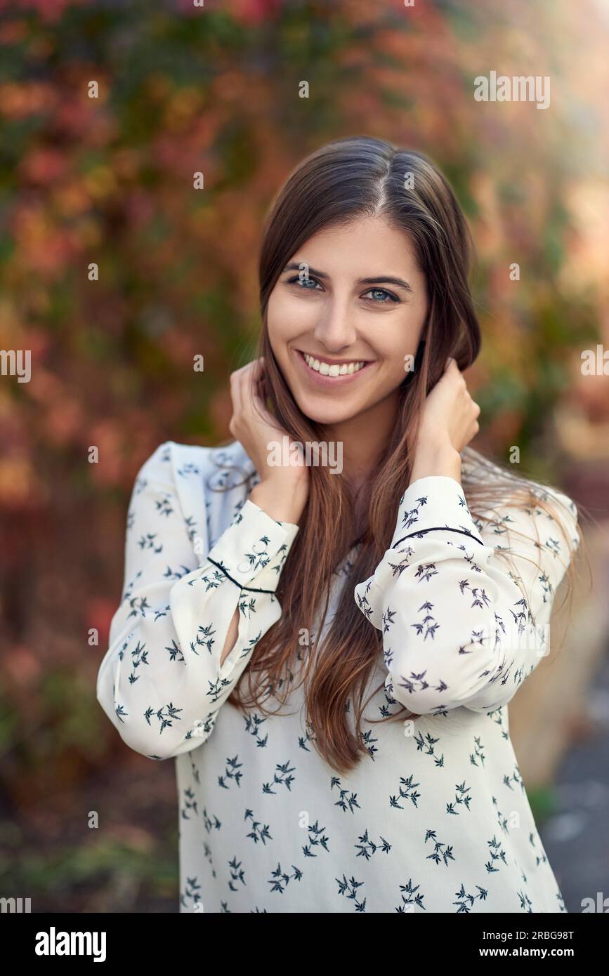 Attraente giovane donna in un colorato cadere o autunno street lateralmente permanente per la fotocamera con la sua mano alla sua guancia sorridendo verso la telecamera Foto Stock