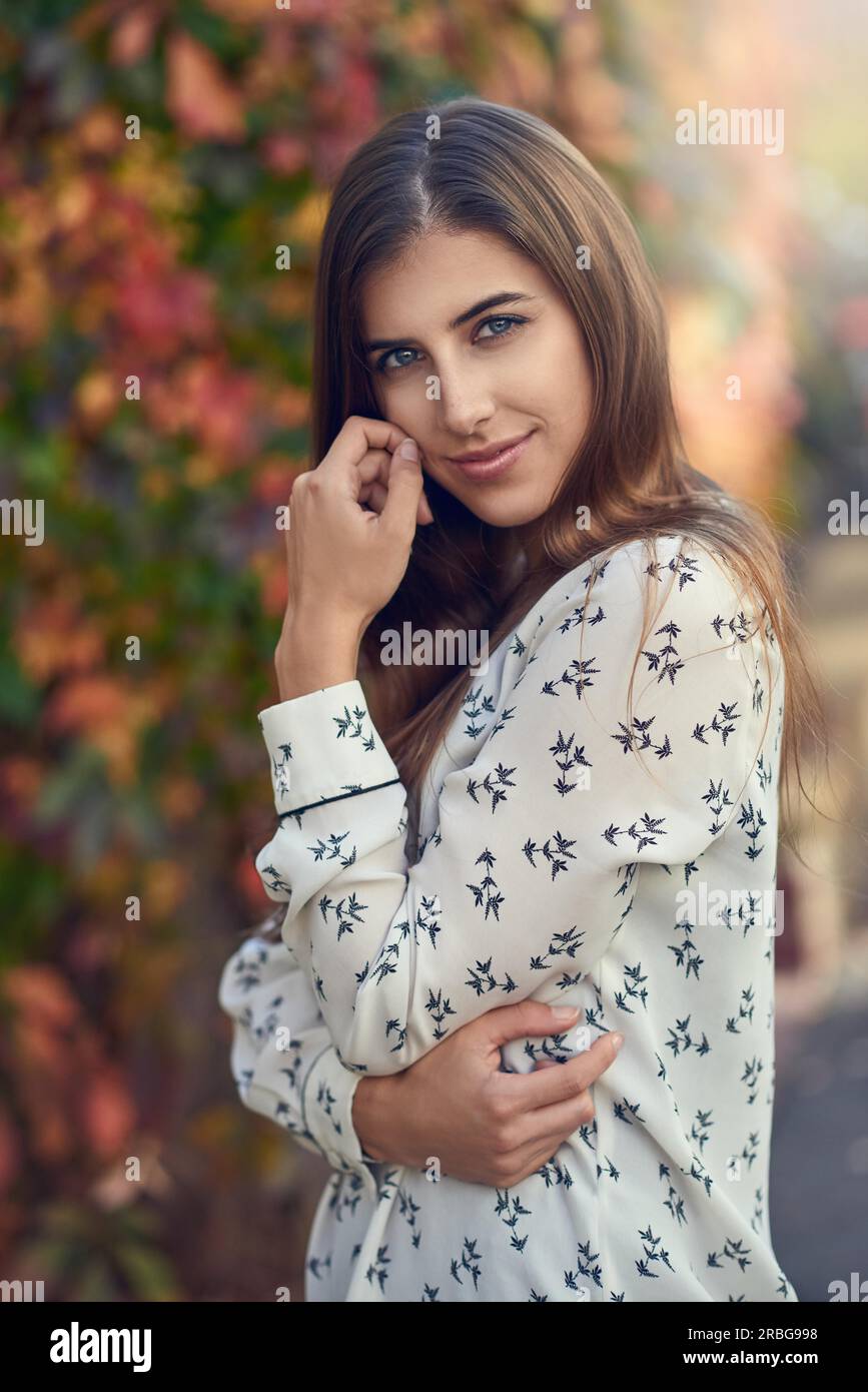 Attraente giovane donna in un colorato cadere o autunno street lateralmente permanente per la fotocamera con la sua mano alla sua guancia sorridendo verso la telecamera Foto Stock