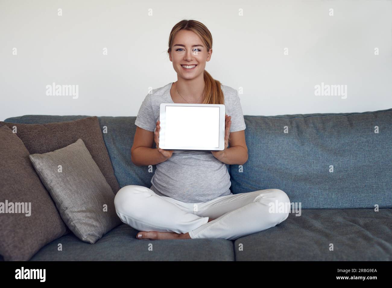Sorridente attraente vivace giovane donna incinta seduta zampe trasversale su un divano a casa con in mano una tavoletta con schermo vuoto e spazio di copia Foto Stock