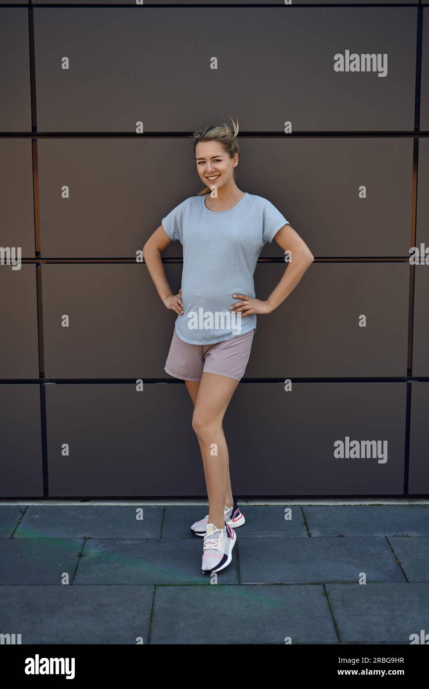 Montare athletic giovane donna incinta in abbigliamento sportivo che pongono di fronte a un edificio commerciale con le mani sui fianchi di sorridere alla telecamera Foto Stock