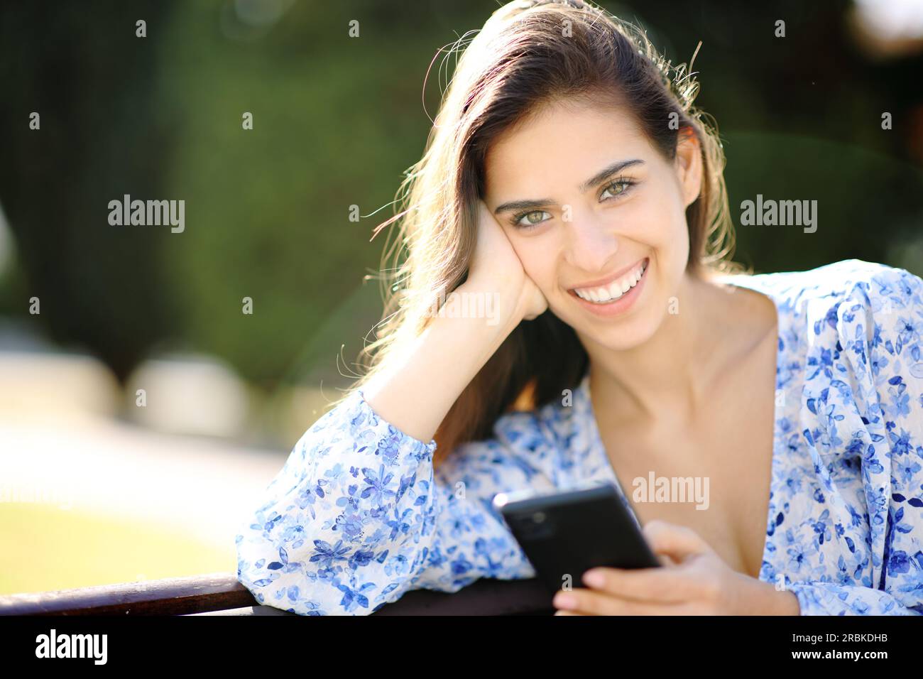 Bella donna felice che posa guardando la fotocamera mentre tiene il telefono in un parco Foto Stock