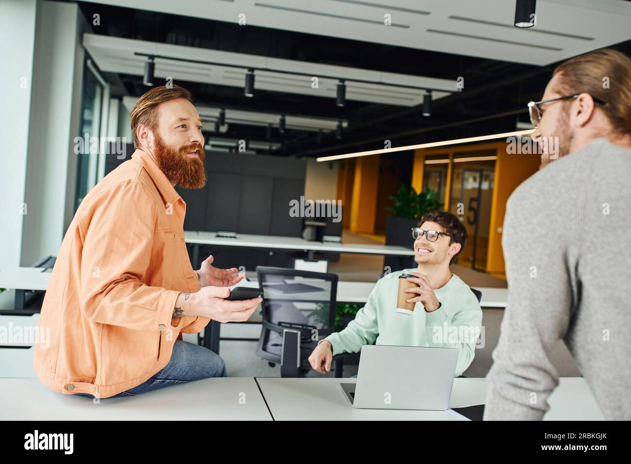 uomo barbuto ispirato che tiene uno smartphone e discute di un nuovo progetto aziendale con i colleghi vicino a un computer portatile sulla scrivania di lavoro in un moderno ufficio di coworking, Foto Stock