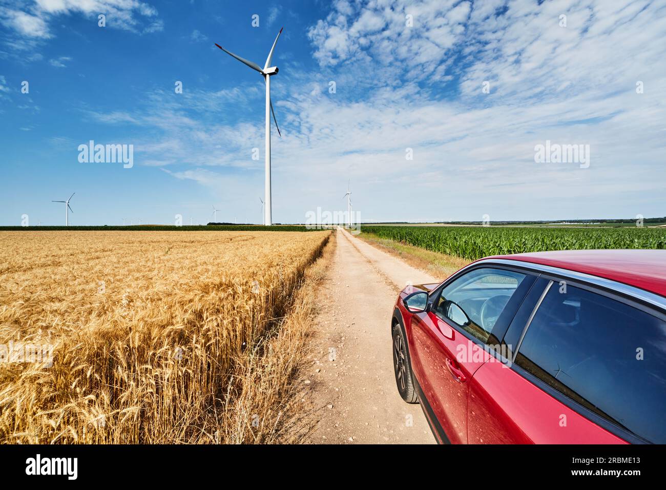 Paesaggio rurale con turbine eoliche, strada di campagna tra campi coltivati e auto rossa Foto Stock