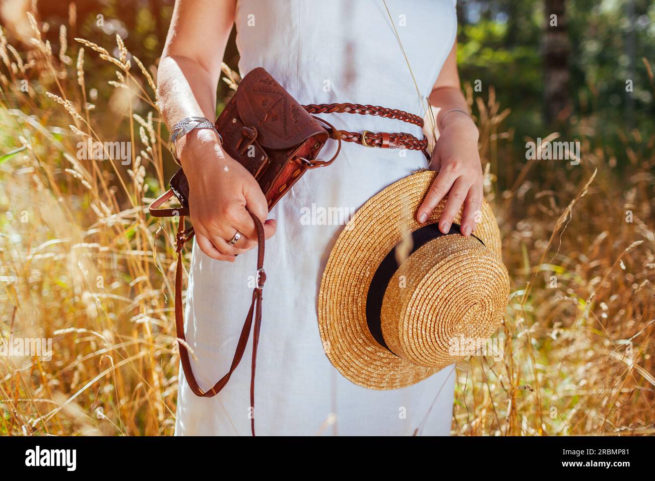 Primo piano della borsa in pelle boho marrone e del cappello di paglia. Donna elegante che indossa abiti bianchi, gioielli, che tiene una borsa all'aperto. bo rustico femminile alla moda Foto Stock