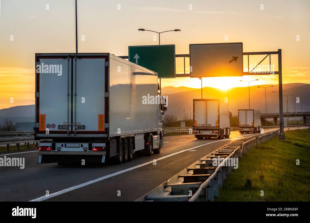 Vista angolare posteriore dei camion da carico per la consegna sulla strada che si dirige verso ovest con il tramonto sullo sfondo. Consegna rapida, logistica merci e spedizioni merci Foto Stock