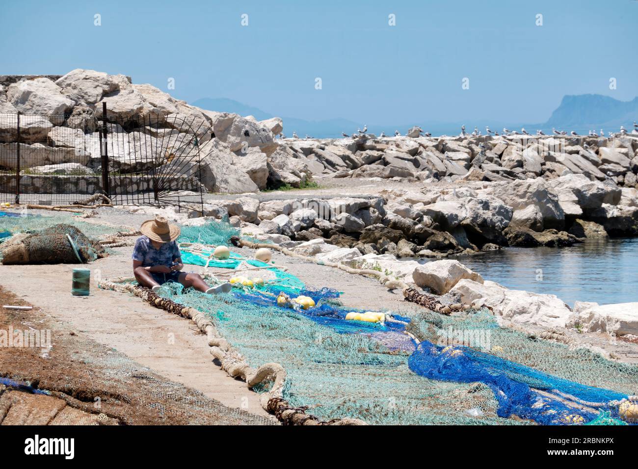 Un pescatore che ripara le sue reti da pesca sul lato del porto di pesca. Il porto si trova a Estepona, Spagna, con Gibilterra sullo sfondo Foto Stock