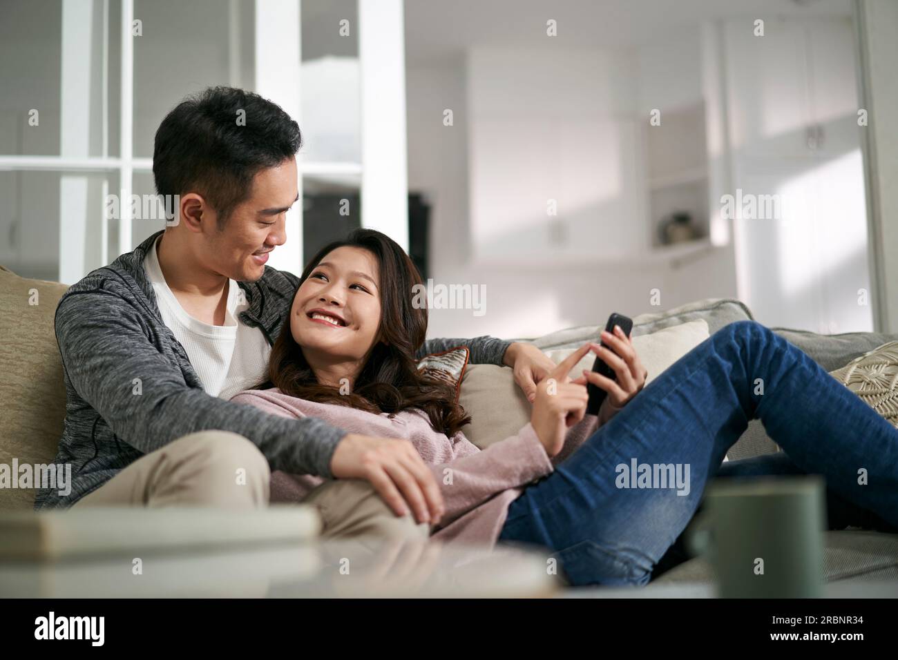 felice giovane coppia asiatica seduta sul divano della famiglia a casa guardando insieme le foto del cellulare Foto Stock