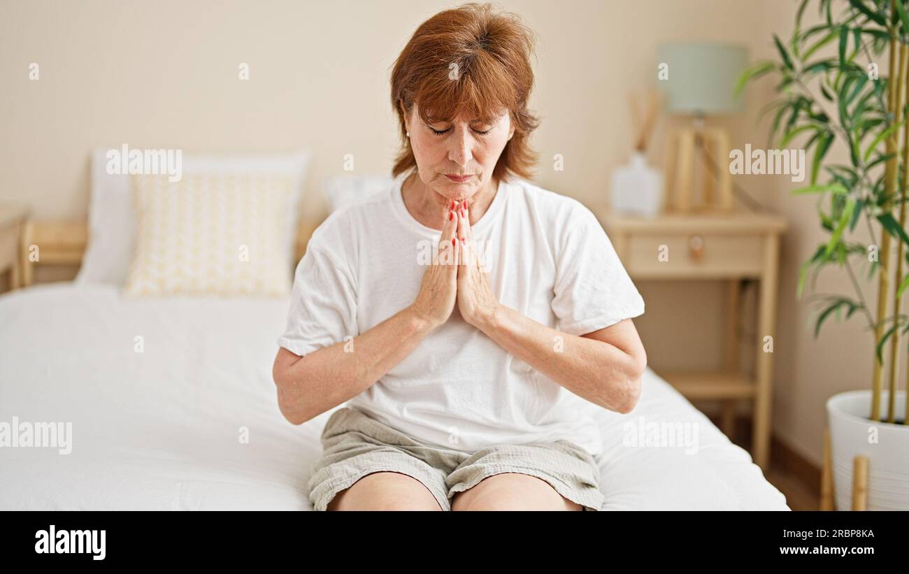 Donna di mezza età seduta sul letto che prega in camera da letto Foto Stock