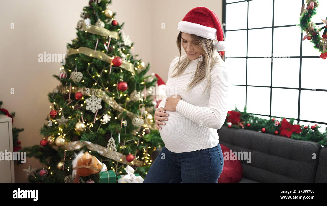 Giovane donna incinta che indossa un cappello di natale che tocca la pancia a casa Foto Stock