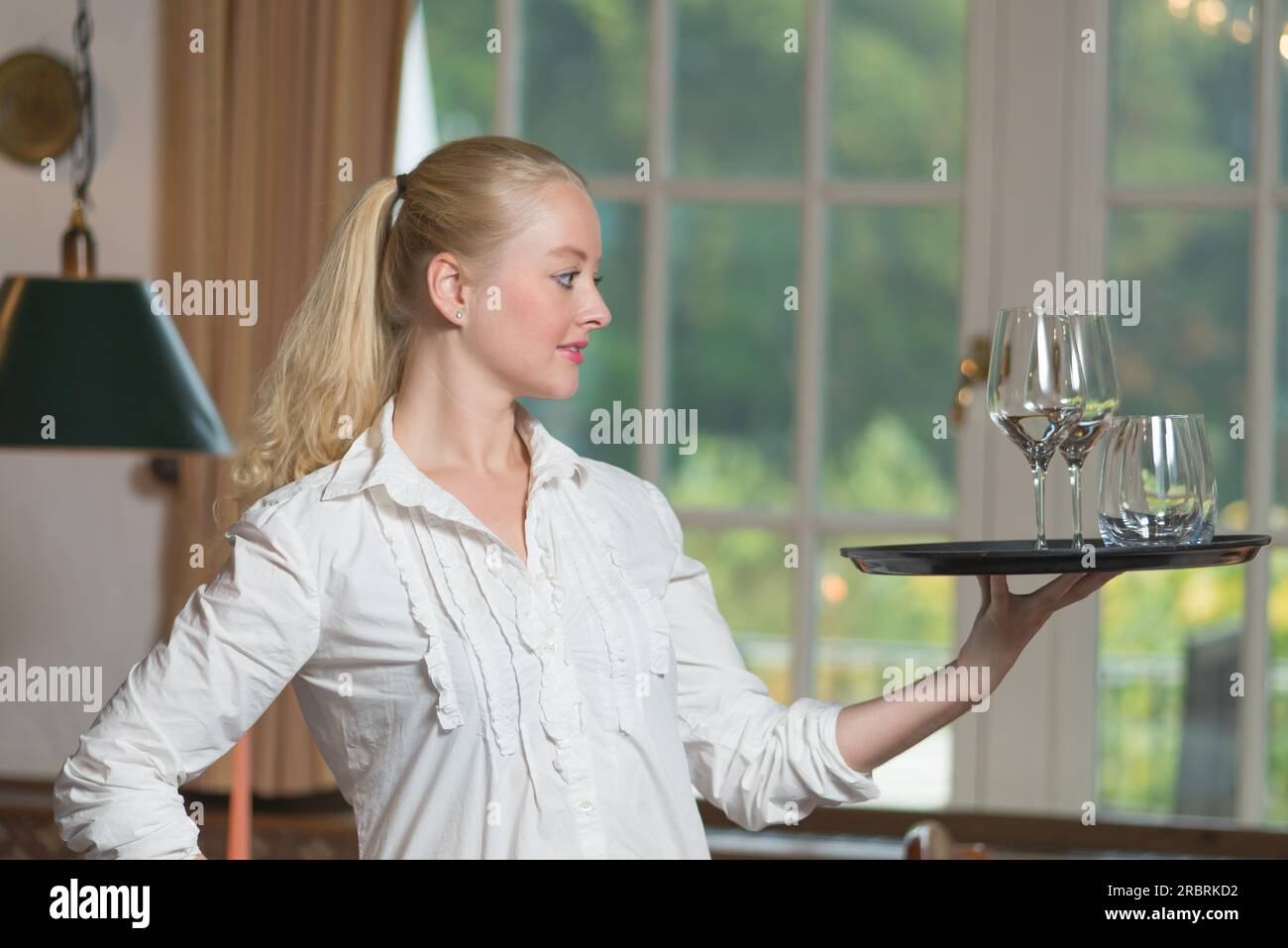 Elegante e bella giovane donna che serve bevande bilanciando con grazia il vassoio con bicchieri di vino bianco sulla punta delle dita in piedi di profilo con un Foto Stock