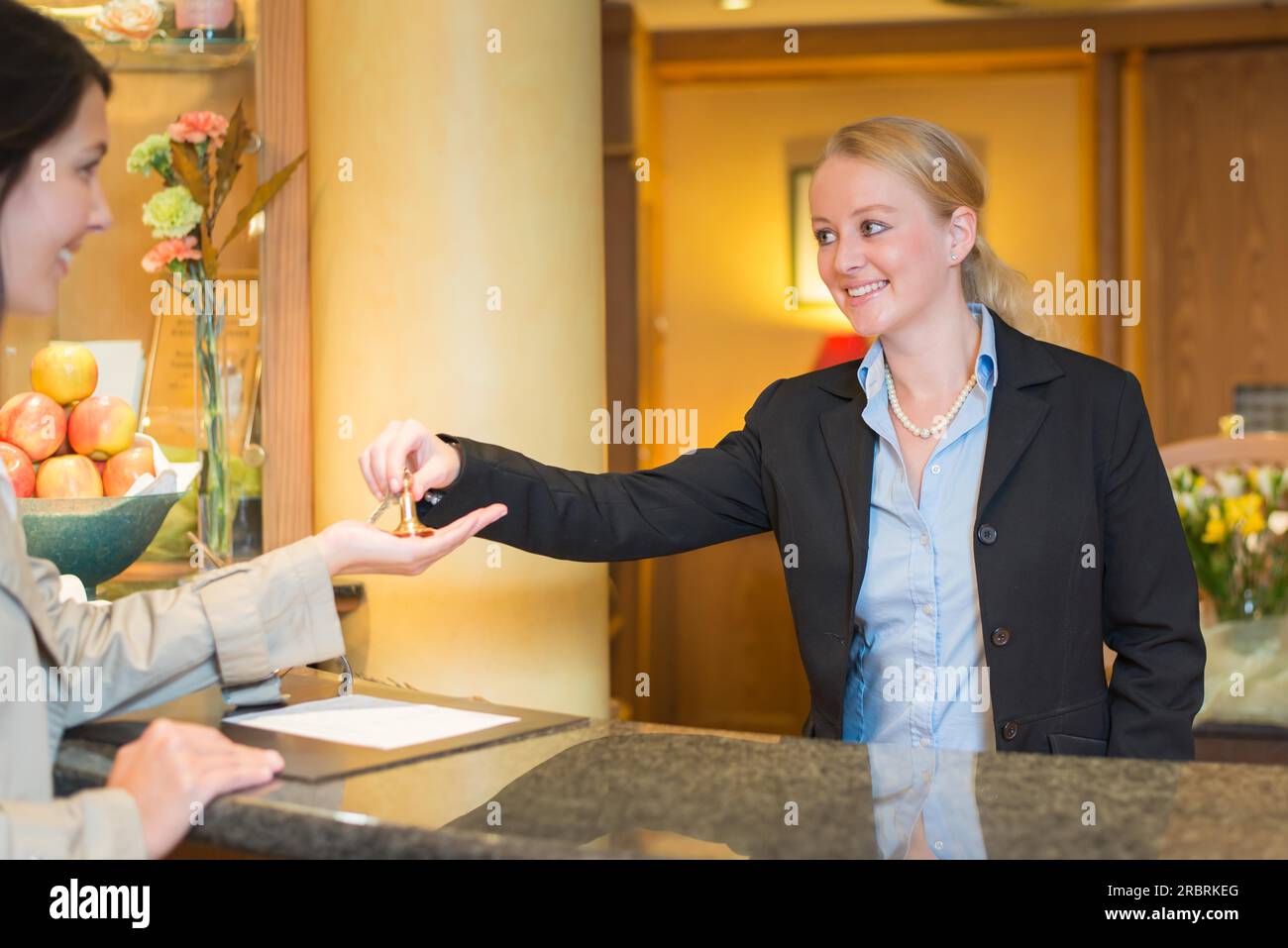 La receptionist dell'hotel sorridente e cordiale si trova dietro il banco del servizio nella hall di un hotel, prenotando con una cliente donna che le consegna le chiavi della camera Foto Stock