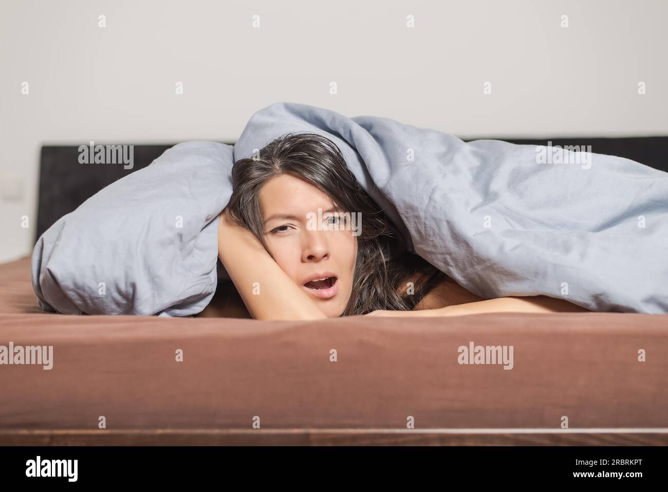 Giovane donna stanca che sbadiglia sotto un piumone mentre trascorre una giornata pigra rilassandosi a casa Foto Stock
