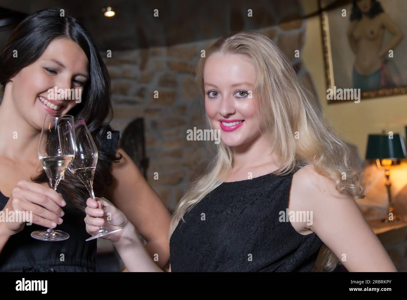 Due eleganti e sofisticate giovani donne indossate in elegante abito nero da sera che celebrano insieme bevendo flute di champagne in una biblioteca dell'hotel o nel lusso Foto Stock