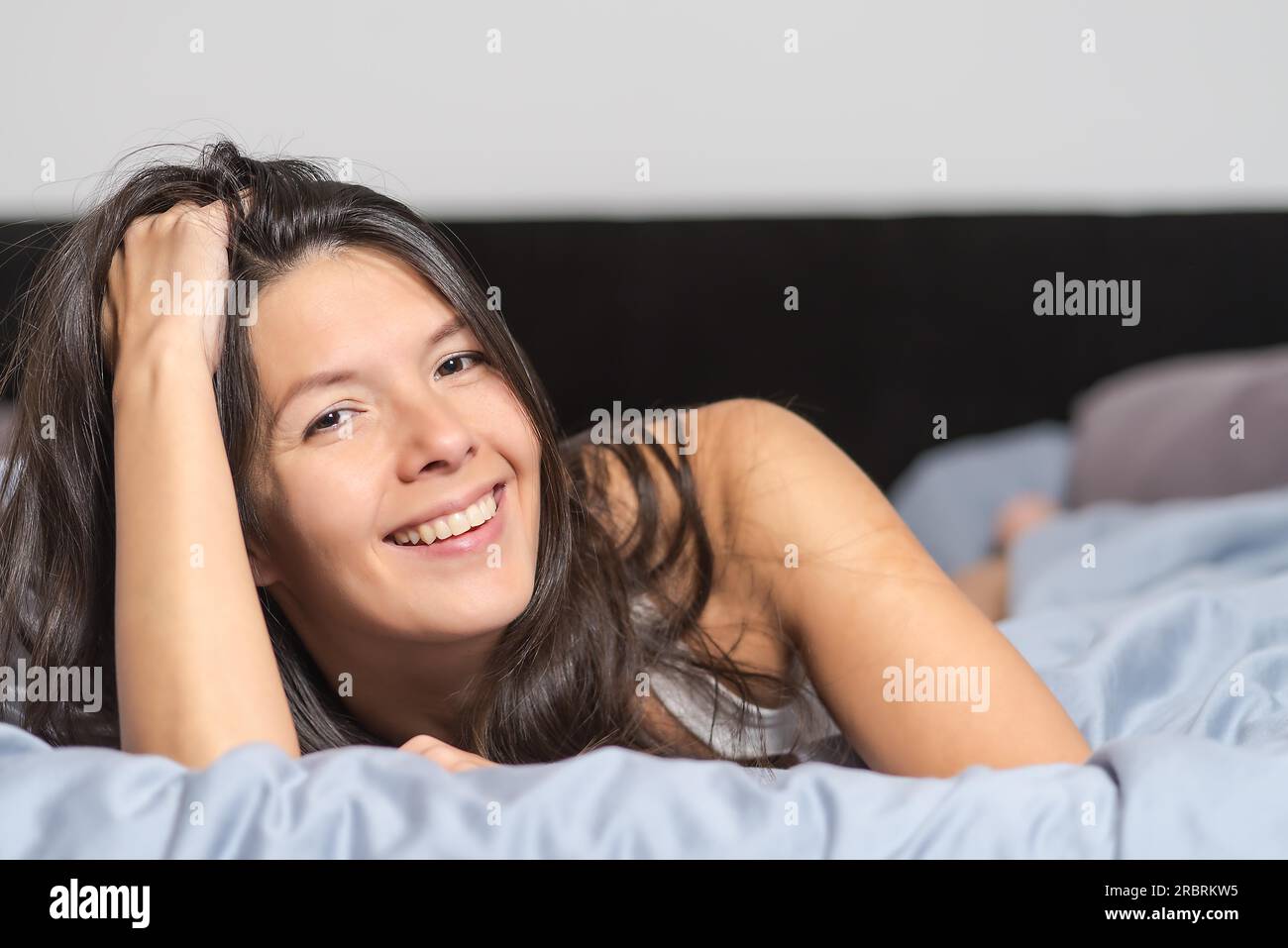 Sorridente attraente giovane donna godendo un giorno pigro giacente sul suo stomaco coccole su un caldo piumone e guardando la telecamera con un sorriso Foto Stock