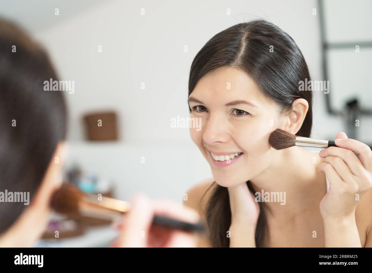 Attraente donna sorridente con i suoi lunghi capelli bruna che applica il trucco in uno specchio inclinato in avanti per vedere meglio mentre applica più blu con un grande Foto Stock