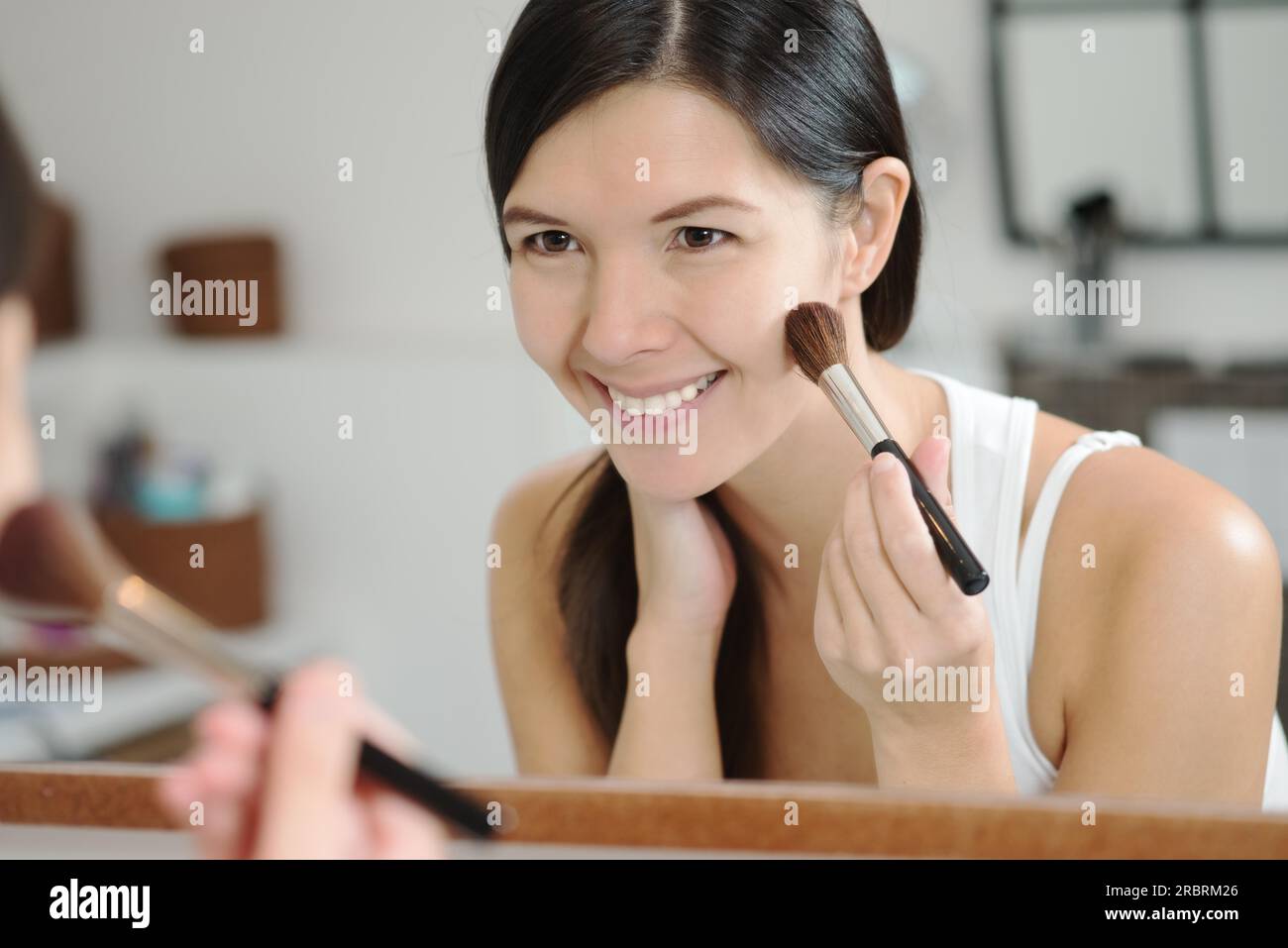 Attraente donna sorridente con i suoi lunghi capelli bruna che applica il trucco in uno specchio inclinato in avanti per vedere meglio mentre applica più blu con un grande Foto Stock