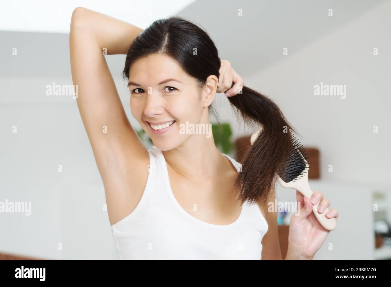 Donna sorridente e attraente che si spazzolava i suoi lunghi capelli bruna in bagno la mattina, tenendola in una mano in una coda di cavallo mentre lei correva la spazzola Foto Stock