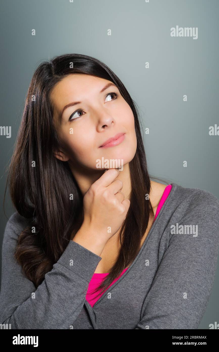 Attraente donna creativa che usa la sua immaginazione seduta in un pensiero profondo con la mano sul mento guardando in alto nell'aria con un sorriso contento Foto Stock