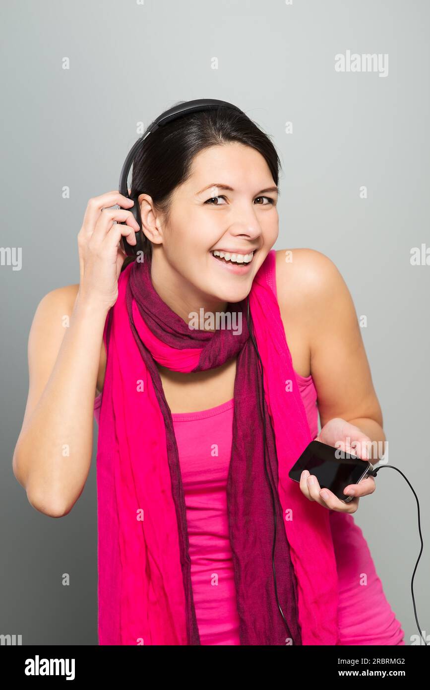 Bella donna vivace ascoltando musica su cuffie collegate al dispositivo elettronico del dispositivo di memorizzazione come essa si erge ridere per la fotocamera Foto Stock