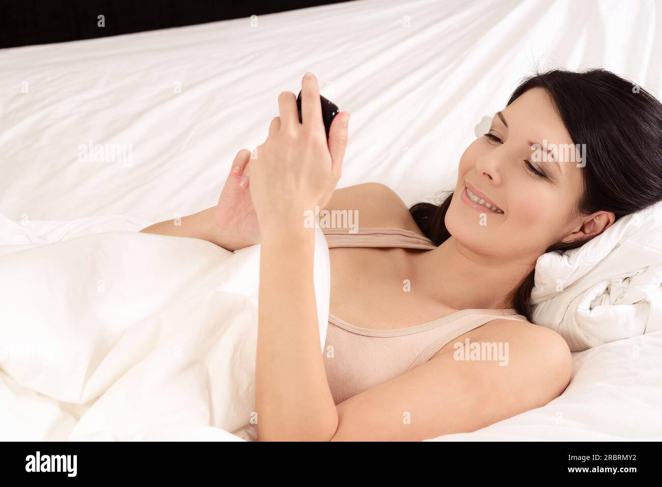 Attraente giovane donna giaceva a letto la lettura su sms sul suo telefono cellulare sorridente con piacere al messaggio, closeup vista laterale Foto Stock
