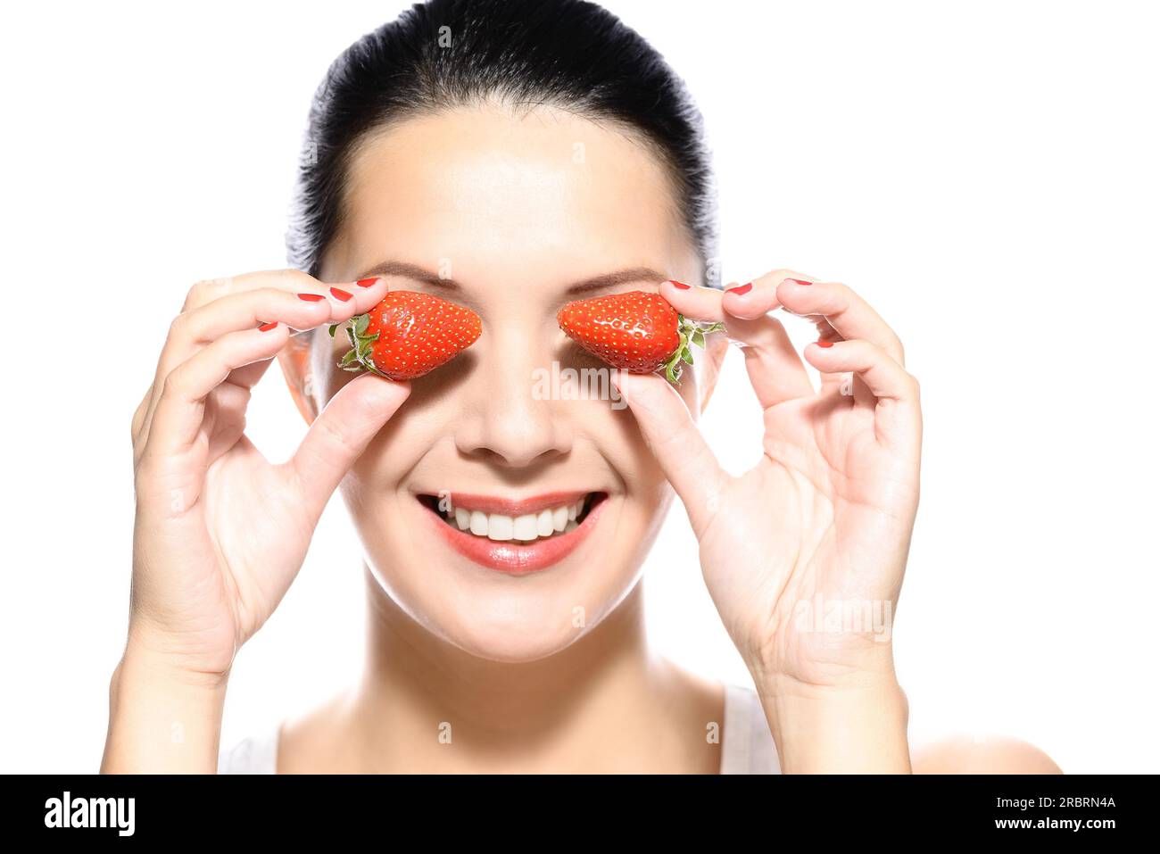 Bella giovane donna con un bel sorriso caloroso e sana la pelle abbronzata tenendo due ripe rosso Fragole ai lati dei suoi occhi, isolato su bianco Foto Stock