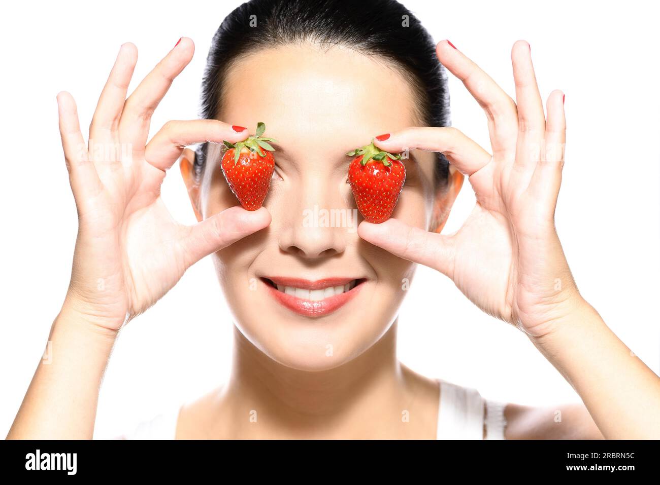 Bella giovane donna con un bel sorriso caloroso e sana la pelle abbronzata tenendo due ripe rosso Fragole ai lati dei suoi occhi, isolato su bianco Foto Stock