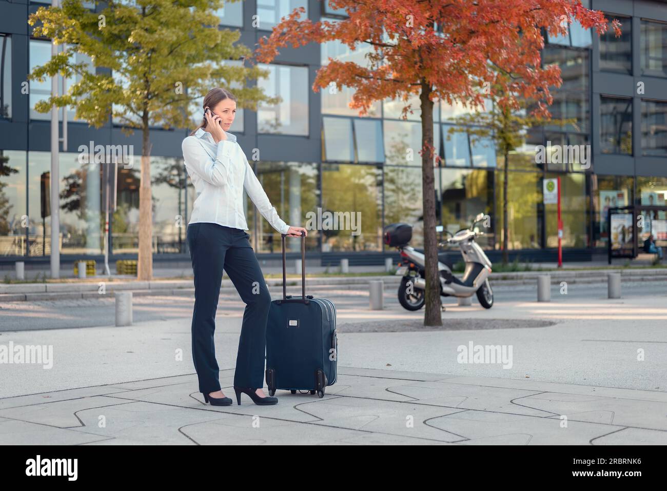 Giovane donna d'affari attiva che parla al telefono cellulare mentre aspetta all'aperto accanto alla custodia del trolley, pronta a viaggiare Foto Stock