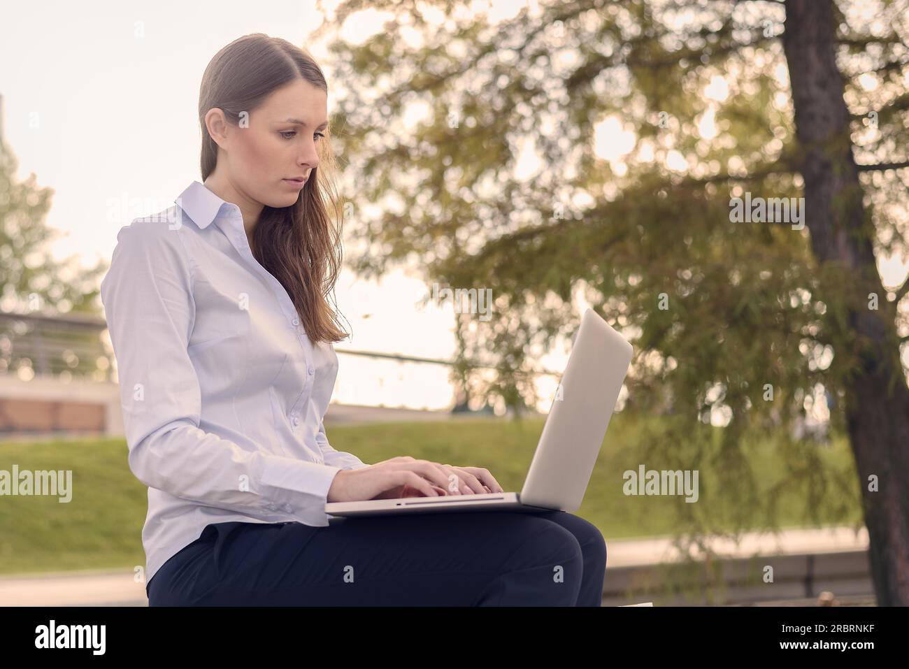 Attraente giovane donna di indossare abiti formali mentre si lavora all'aperto su un computer portatile con una connessione wireless a internet, in un bel giorno di estate Foto Stock