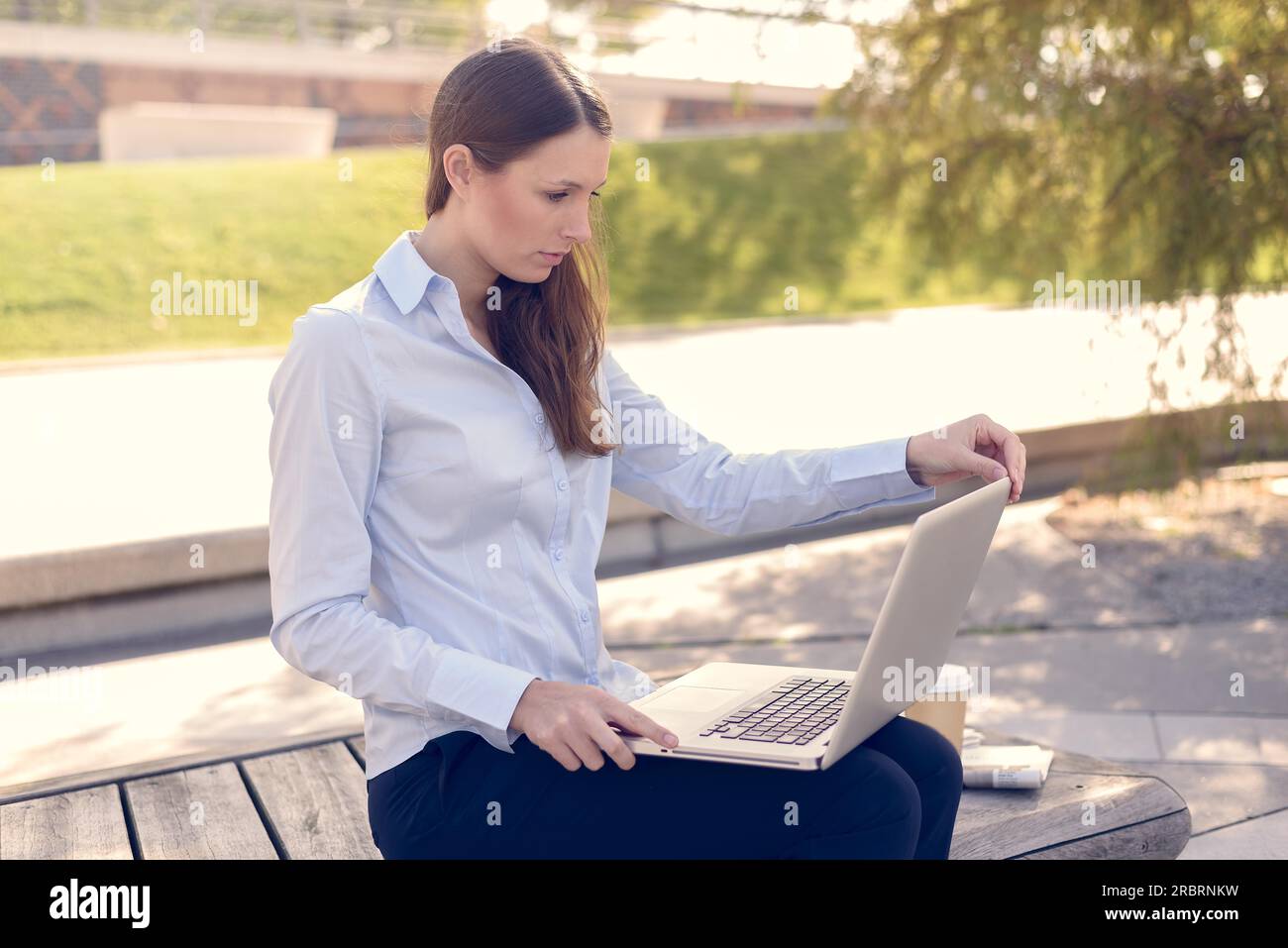 Attraente giovane donna di indossare abiti formali mentre si lavora all'aperto su un computer portatile con una connessione wireless a internet, in un bel giorno di estate Foto Stock