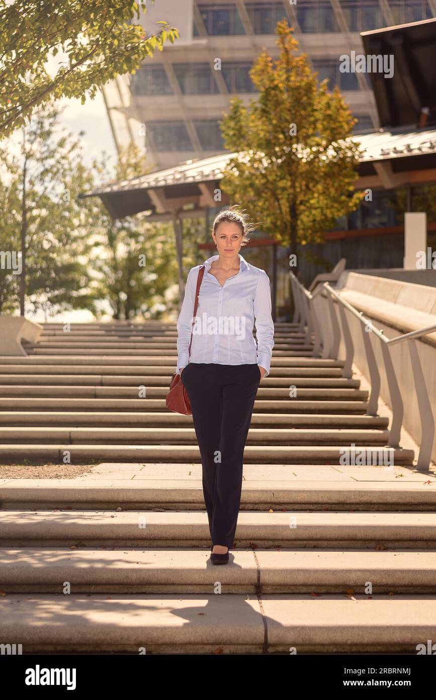 Snello ed elegante giovane donna che porta una borsetta scendendo di un volo di open-air scale di cemento in città con moderni edifici commerciali dietro di lei Foto Stock