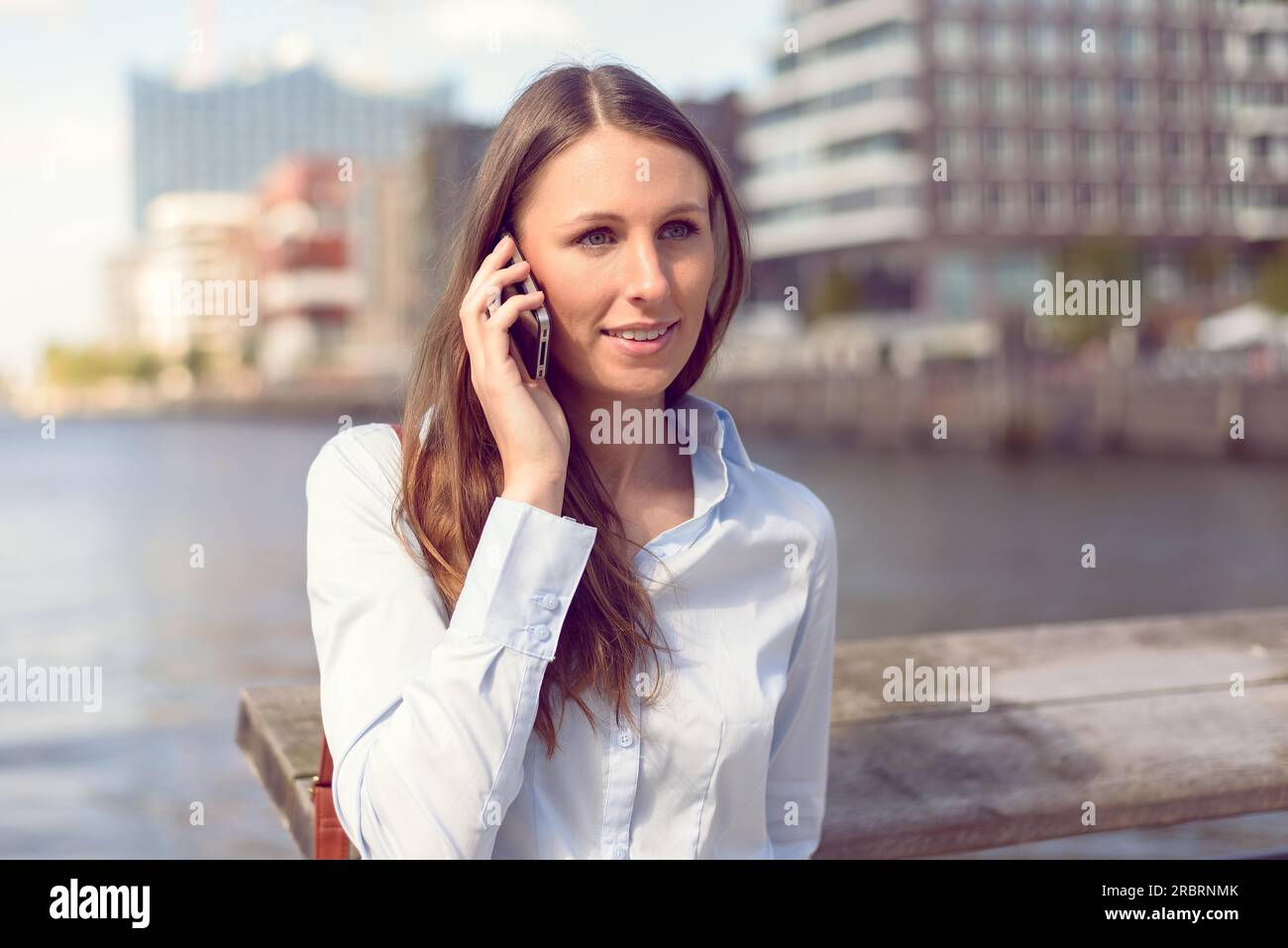Donna attraente che riceve una chiamata sul suo telefono cellulare in piedi all'aperto su uno sfondo fluviale sorridendo davanti alla fotocamera mentre ascolta il Foto Stock