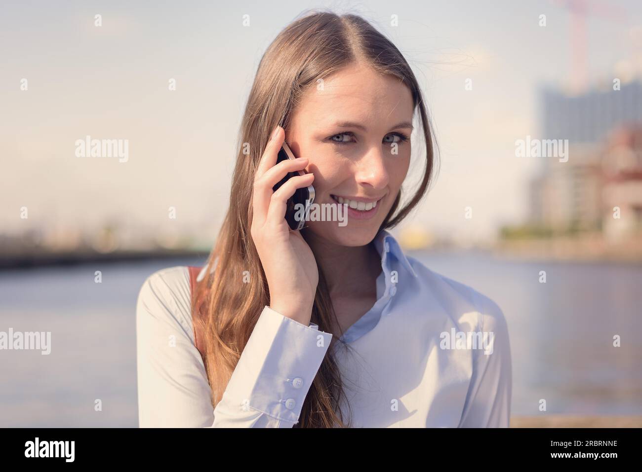 Donna attraente che riceve una chiamata sul suo telefono cellulare in piedi all'aperto su uno sfondo fluviale sorridendo davanti alla fotocamera mentre ascolta il Foto Stock