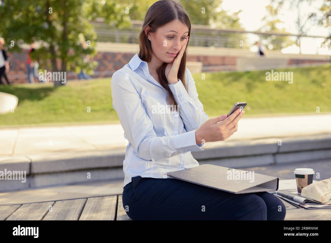 Giovane donna attraente e snella seduta all'aperto in un parco urbano mentre legge un sms sul suo cellulare con un aspetto preoccupato e la mano sul volto Foto Stock