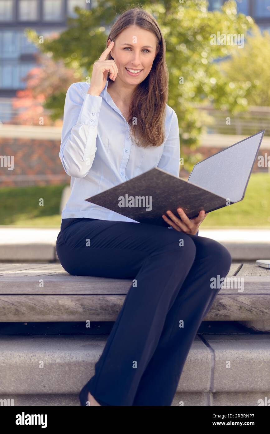 Sorridente attraente imprenditrice avente un'idea durante la lettura di una cartella di business in un parco Foto Stock
