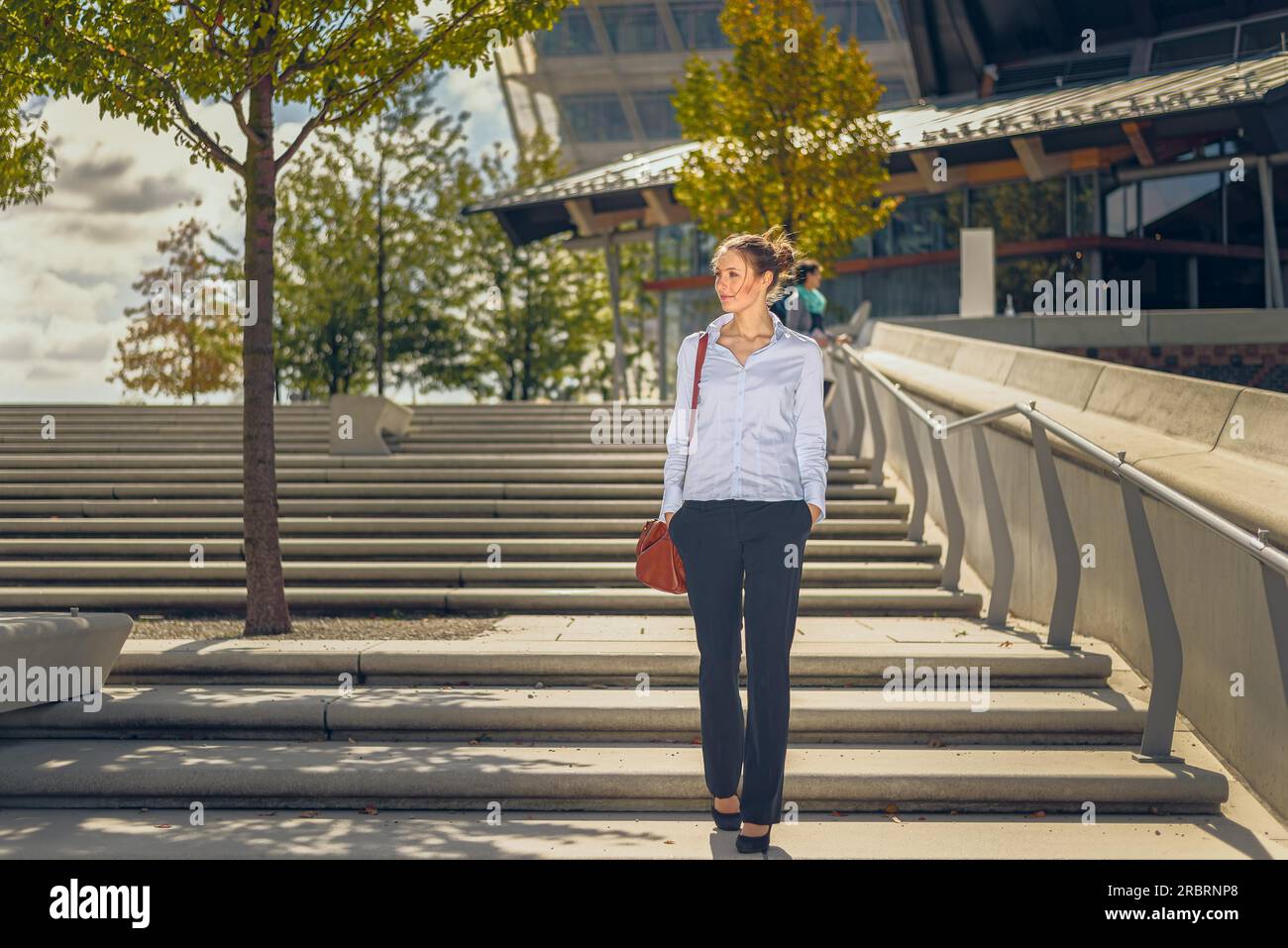 Snello ed elegante giovane donna che porta una borsetta scendendo di un volo di open-air scale di cemento in città con moderni edifici commerciali dietro di lei Foto Stock